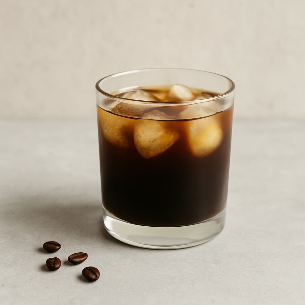 Stiklinė su šalta kava ir ledukais ant šviesaus stalo, šalia keli kavos pupelių grūdai.